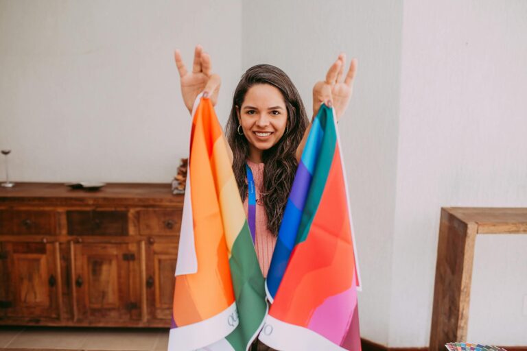 Smiling woman holding colorful rainbow flags, celebrating indoors with joy and pride.